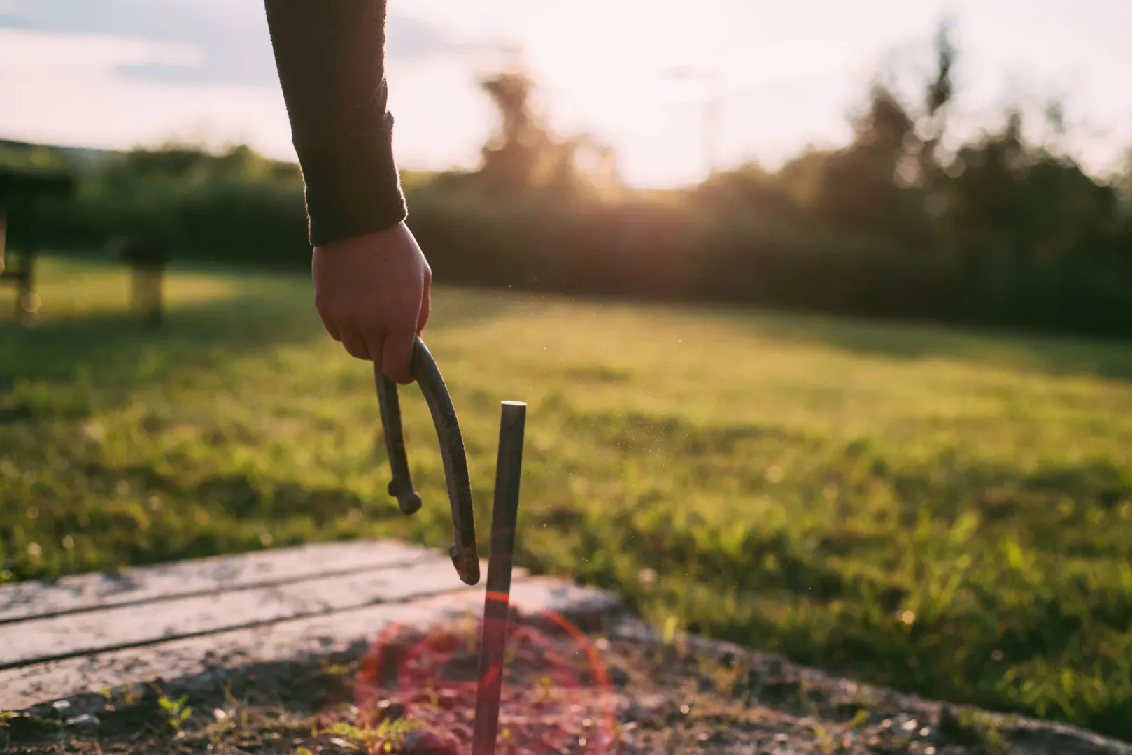A player pitching a horseshoe toward the stake on a grassy backyard court at sunset